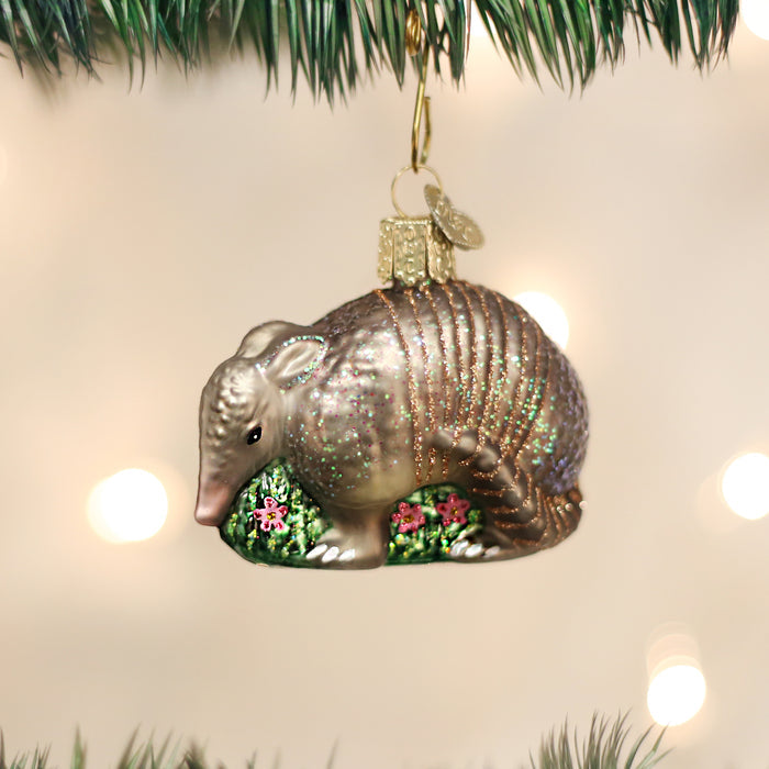 A Christmas tree ornament shaped like a glittery armadillo stands on a green grassy base with small pink flowers, hanging among green pine needles with blurred lights in the background.