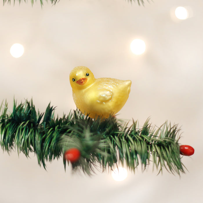 A yellow duck ornament sits on a green pine branch with red berries, against a softly blurred background with glowing white lights.