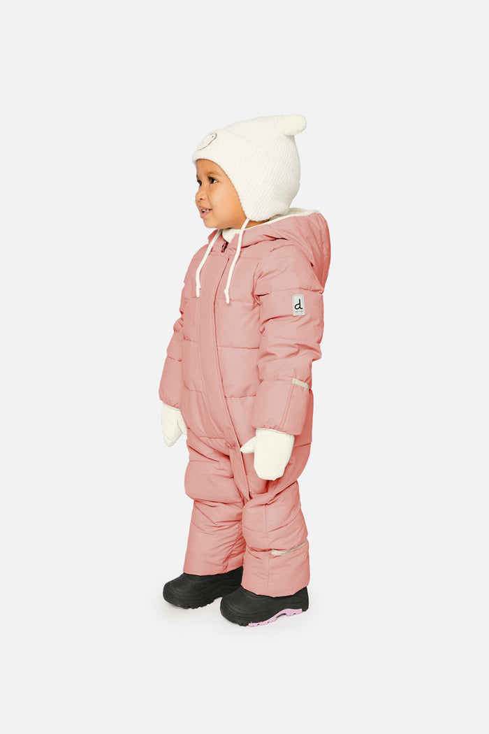 Baby standing in the pale dusty pink Deux par Deux Baby Cloud snowsuit with a cream hat and mittens, side view