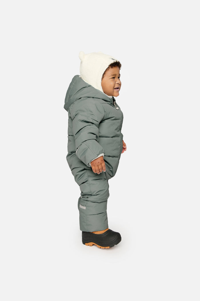 Baby standing in the sage green Deux par Deux Baby Cloud snowsuit, side view