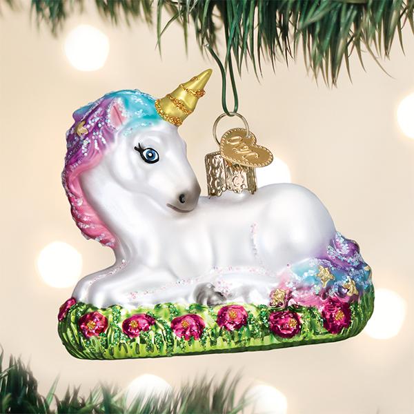A shiny unicorn Christmas ornament with a gold horn, pink and blue mane, and sparkling details rests on green grass with pink flowers, hanging from a Christmas tree branch.