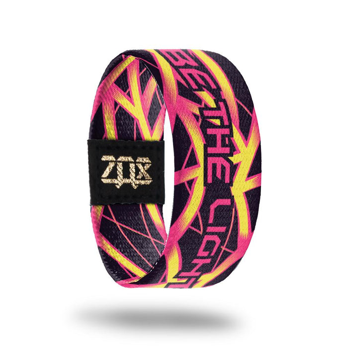 Inside design of Be The Light has a black background with thin pink lines and thicker pink and yellow lines running along the design with no repeating patterns and Be The Light text centered in pink with a black outline around each letter