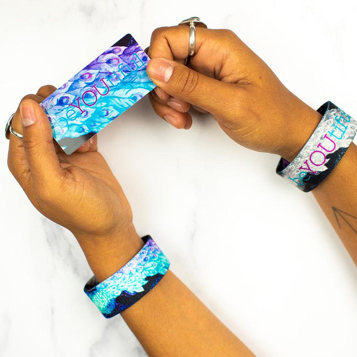 Studio image of hands with rings holding a card that says BeYOUtiful with one BeYOUtiful wristband on each wrist