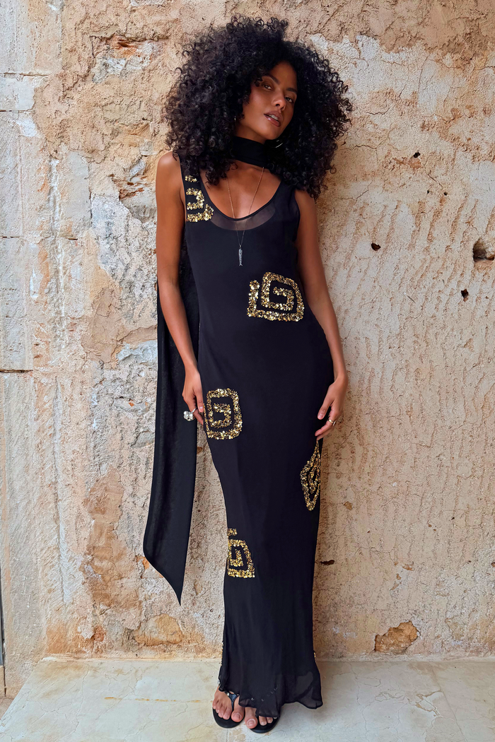 Black and gold Simona petite maxi dress with sequin patterns and a detachable scarf against a stone wall.