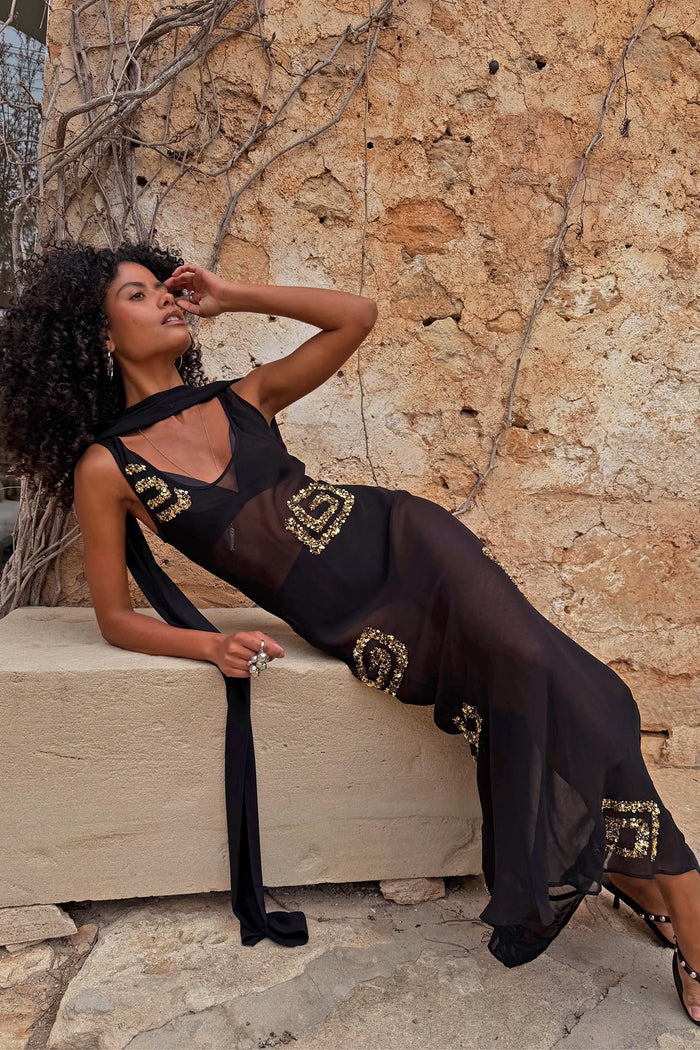 Petite Black & Gold Simona Maxi Dress in sheer mesh with gold sequins and a multi-wear scarf, styled against a stone wall.
