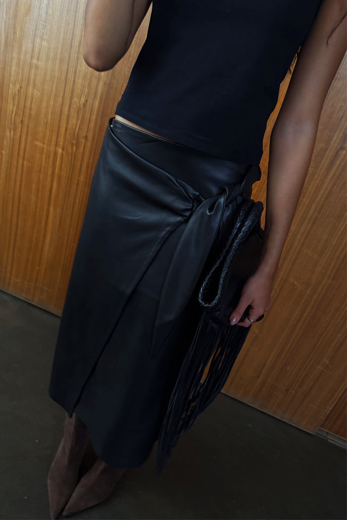 Black vegan leather wrap skirt with a tulip tie detail, styled with a black top and fringe bag.