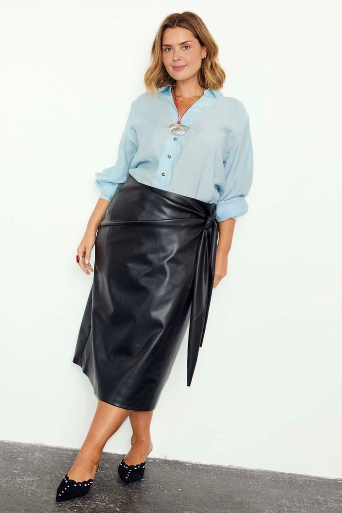 Black vegan leather Jaspre wrap skirt with a tulip-shaped tie, styled with a light blue shirt.