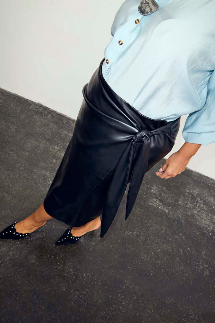 Iconic Black Vegan Leather Wrap Skirt with a tulip shaped tie, styled with a blue shirt and studded black mules.