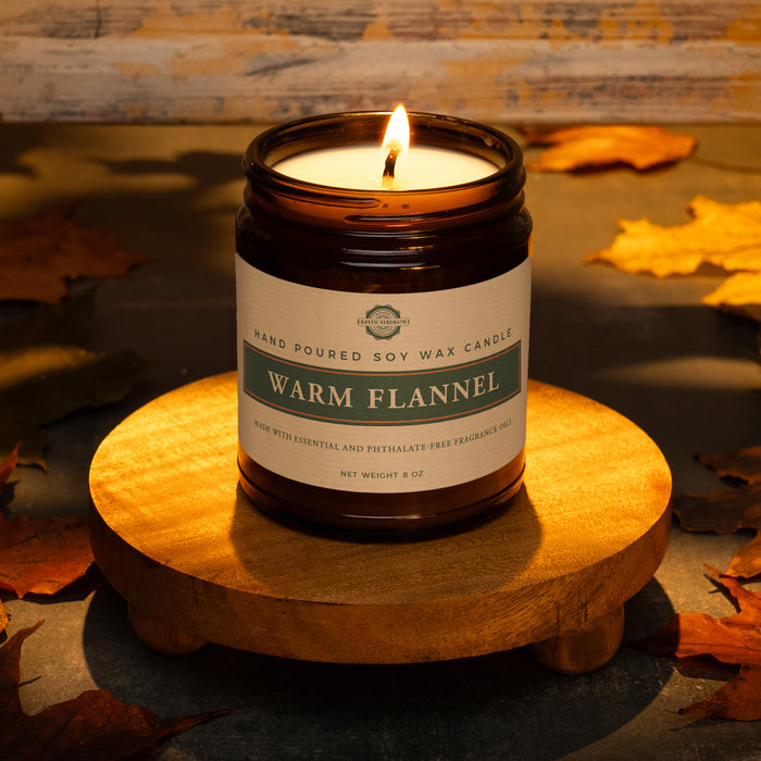 Rustic Strength, Candle, 8oz, Amber Glass, photoshoot image showing a lit candle on a wooden platform set in a rustic, warmly lit setting surrounded by fall leaves. Warm Flannel scent
