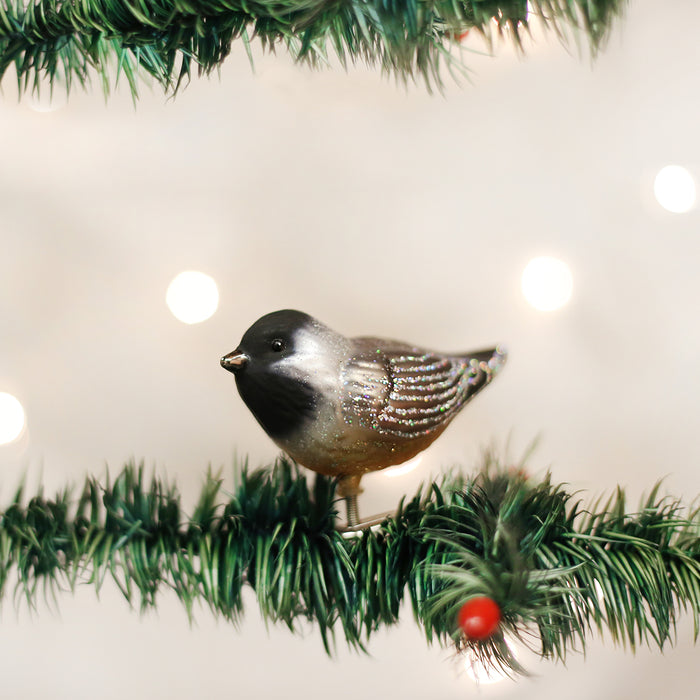 A bird-shaped ornament with black and white coloring sits on a green Christmas tree branch. The background is softly blurred with warm white lights.