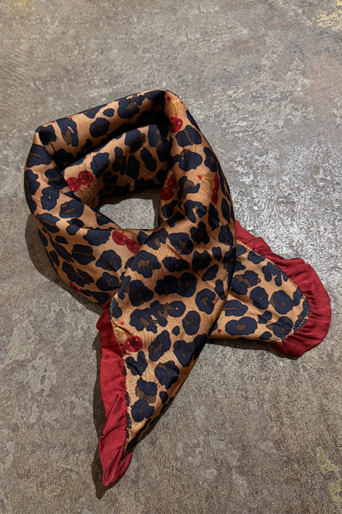 Cherry Leopard Bandana featuring leopard print with red cherries and a deep red ruffle edge on a textured surface.