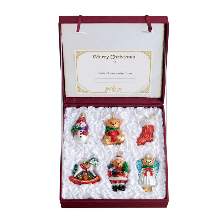 A red gift box with a â€œMerry Christmasâ€_x009d_ card contains seven festive glass ornaments, including teddy bears, a white dog, Santa, a stocking, and a toy rocking horse, arranged on a white satin lining.