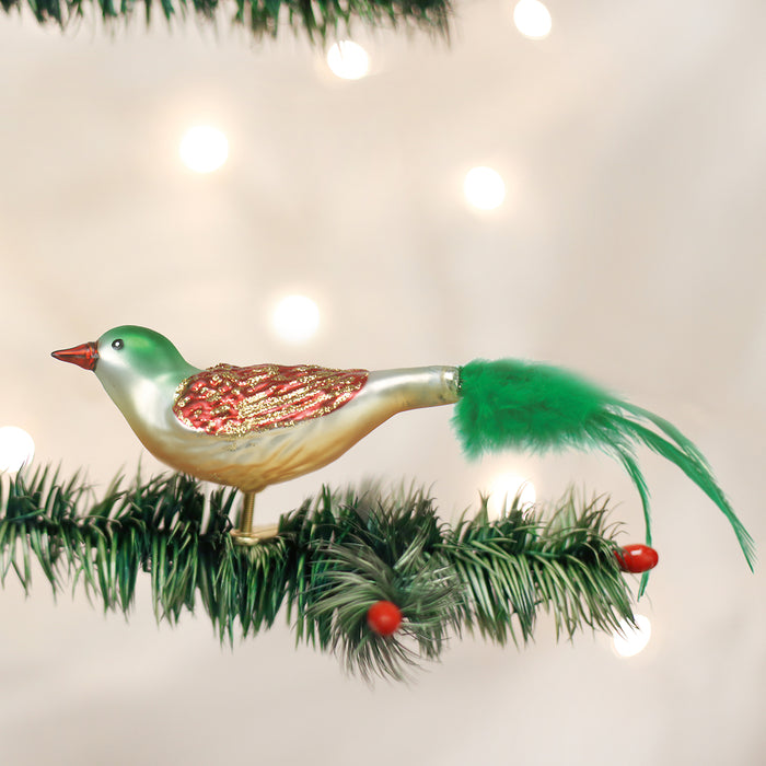 A glass bird ornament with a green head, red beak, gold and red glittery wings, and a long green feather tail clips onto a pine branch decorated with red berries; blurred lights shine in the background.