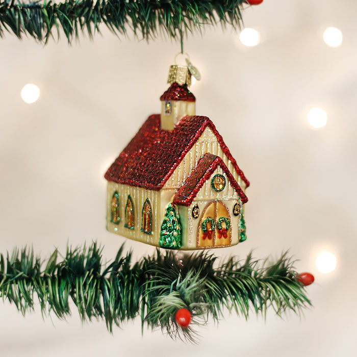 A glittery Christmas church ornament with a red roof hangs on a green tree branch, with warm white lights softly glowing in the blurred background.