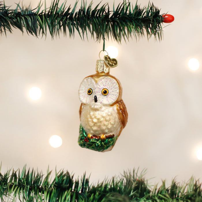 A glass owl ornament with white and brown feathers hangs from a green artificial Christmas tree branch, with a soft, glowing background of out-of-focus lights.