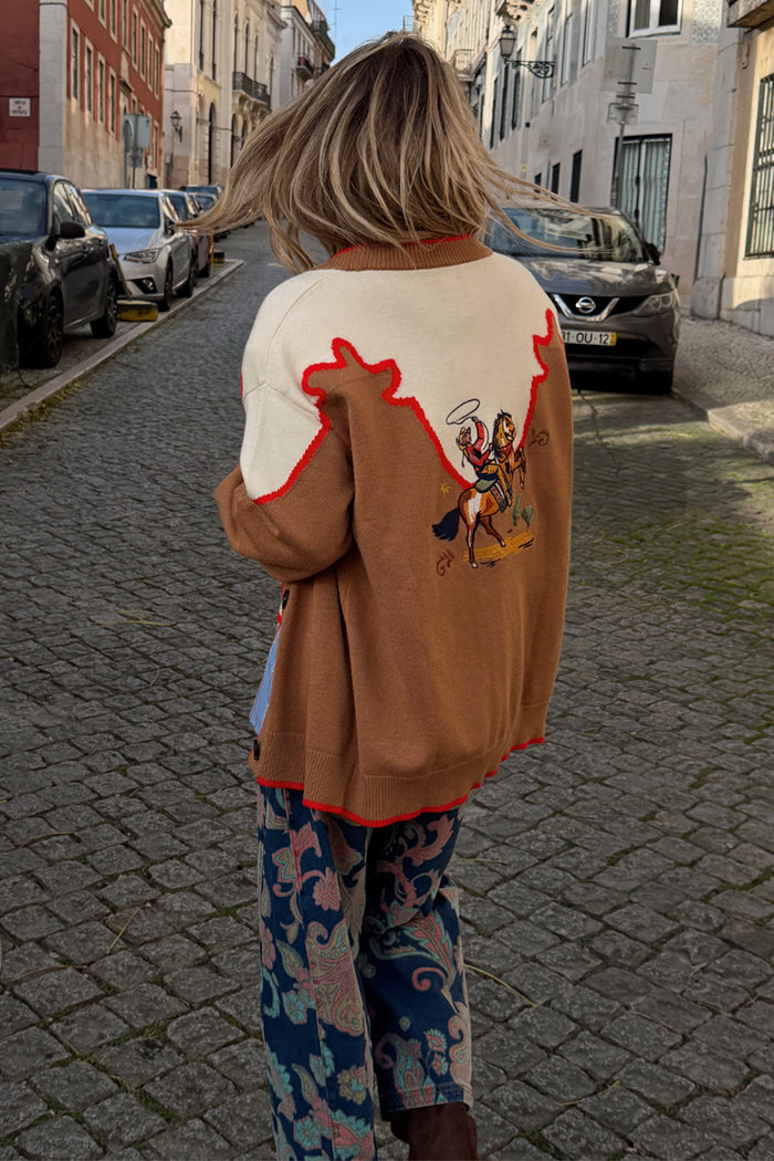 Tan Cowgirl Martha Cardigan with embroidered cowgirl motifs and red stitching, seen on a cobbled London street.