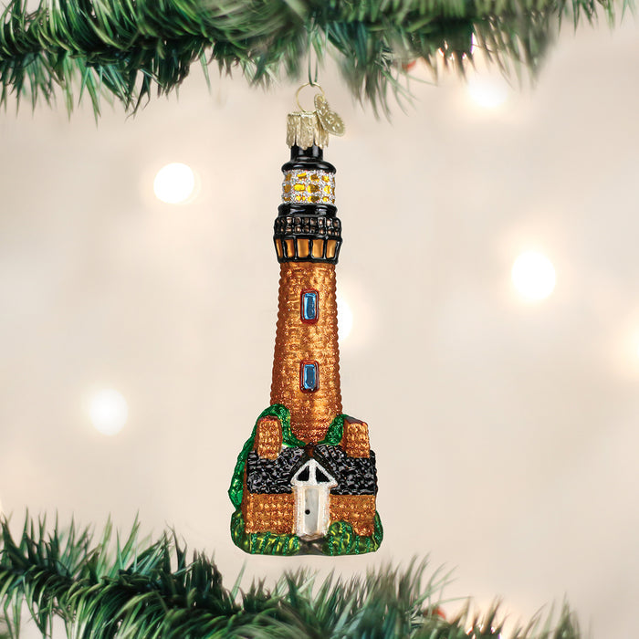 A glass Christmas ornament shaped like a lighthouse with a house at its base, hanging on a green Christmas tree branch, with softly blurred lights in the background.