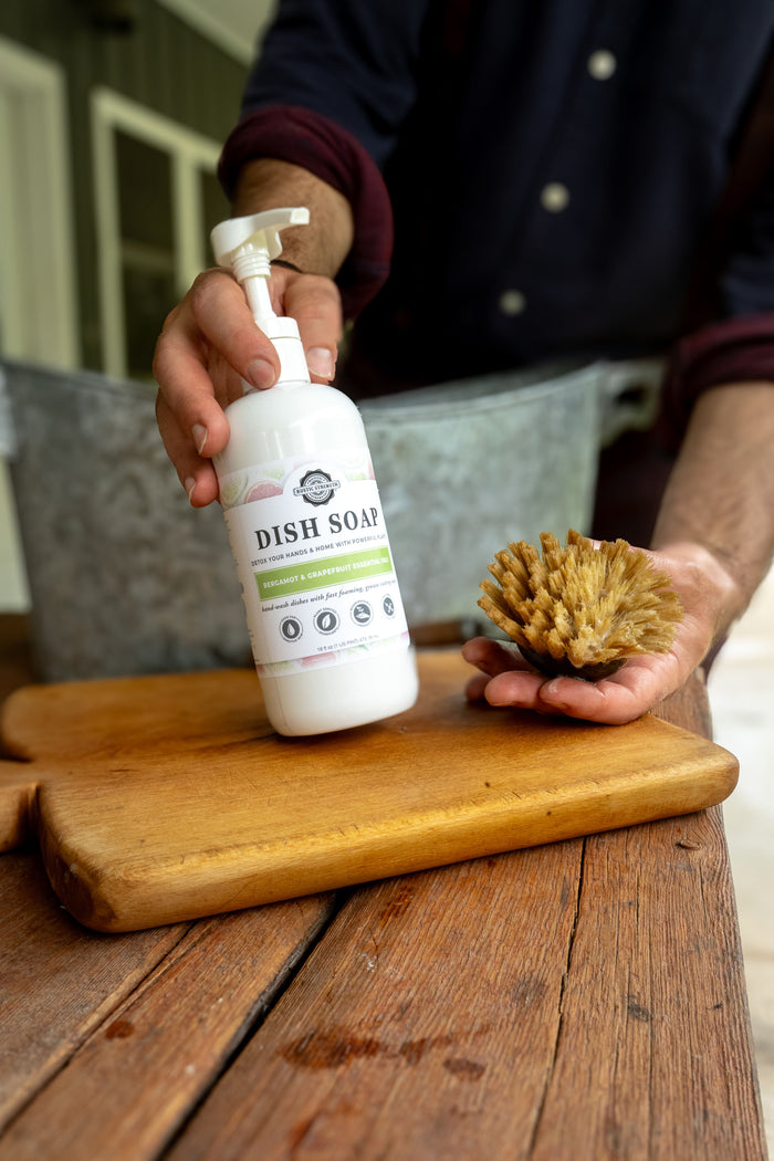 Rustic Strength, Dish Soap, Person holding a Bergamot & Grapefruit dish soap bottle and a brush.