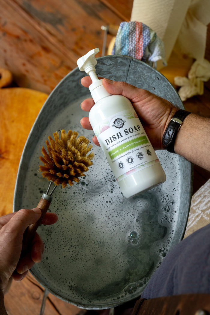 Rustic Strength, Dish Soap, Person holding Bergamot & Grapefruit dish soap and a brush doing dishes.