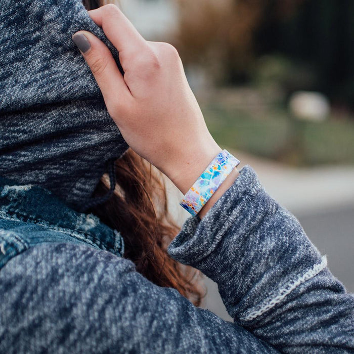 Lifestyle photo of hand grasping hood of jacket wearing dream big single showing outside design with a white geometric design overlaying a blue, purple, and yellow watercolored background