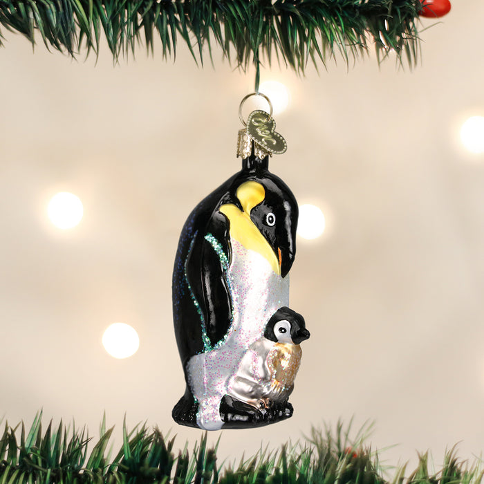 A glass Christmas ornament depicting a penguin with a chick stands out on a green pine tree branch, with warm white lights blurred in the background.