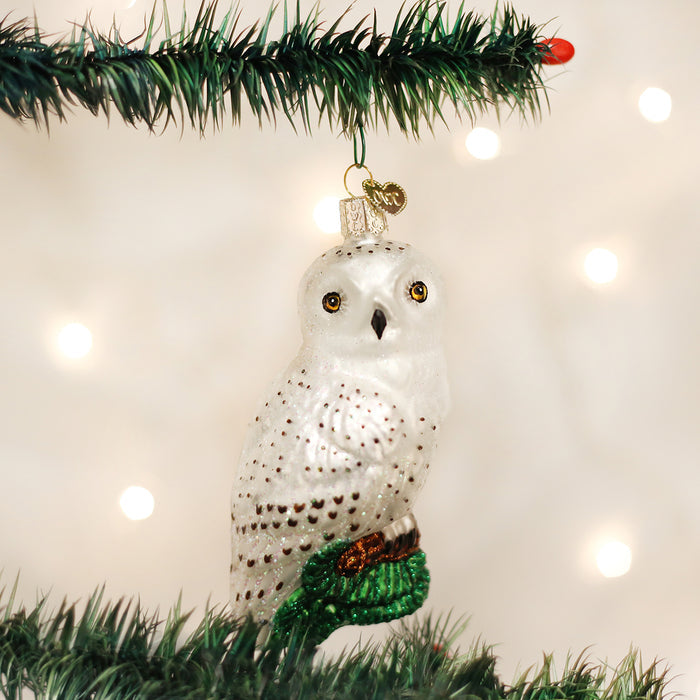 A white owl ornament with gold eyes and black spots hangs from a green Christmas tree branch, with soft, out-of-focus lights in the background.