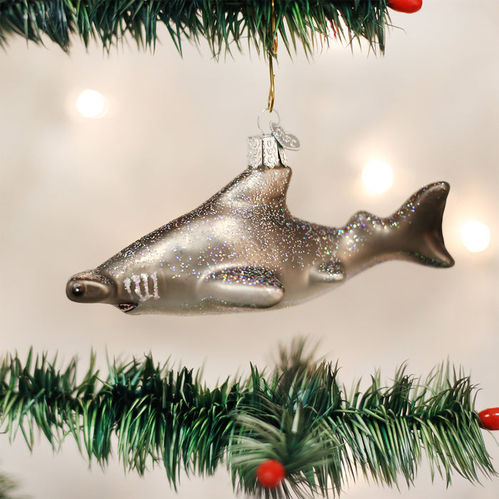 A glittery shark-shaped Christmas ornament hangs from a green, decorated pine tree branch with blurred white lights in the background.