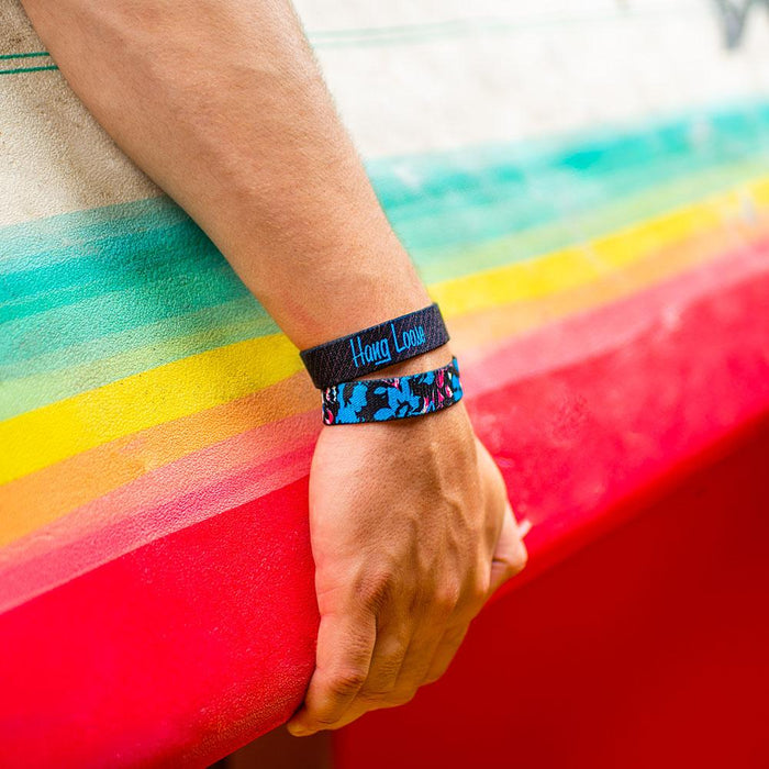 Lifestyle photo of guy holding a surfboard wearing two hang loose singles one with the outside design of neon tropical flower print overlaying black background and below the other showing the inside design of hang loose with italic text hang loose with grey and black striped background