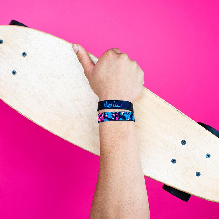 Studio photo of guy holding a skateboard wearing two hang loose singles one with the outside design of neon tropical flower print overlaying black background and below the other showing the inside design of hang loose with italic text hang loose with grey and black striped background