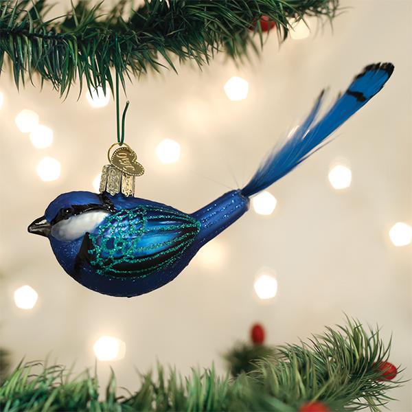 A blue bird ornament with glittery details hangs from a green Christmas tree branch; blurred white lights shine in the background.