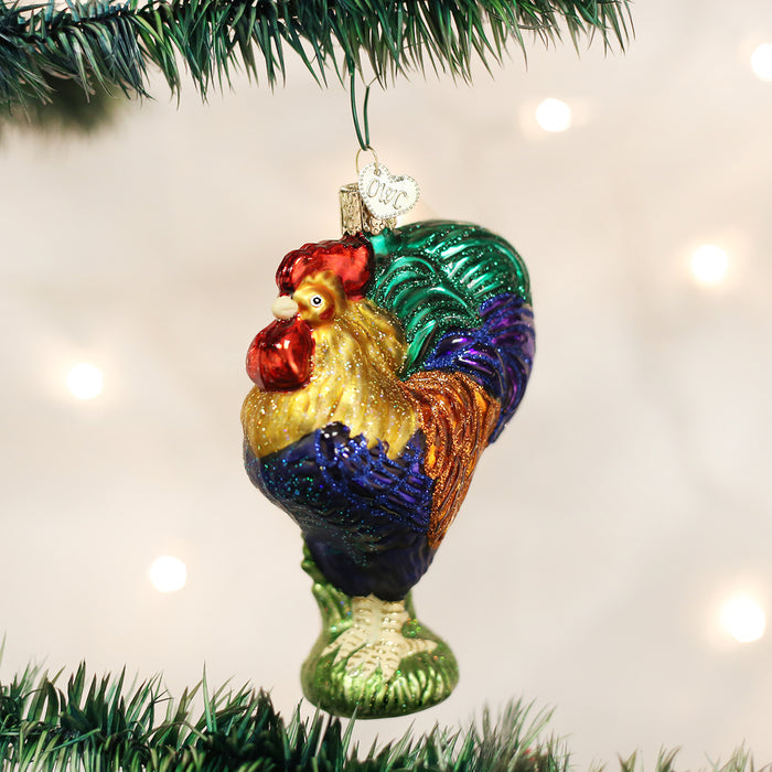 A colorful rooster-shaped glass ornament hangs from a green Christmas tree branch, with soft white lights blurred in the background.