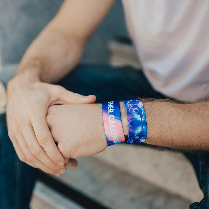 Lifestyle image of a man wearing two Higher Calling straps. One shows the outside design while the other shows the name on the inside. He is holding his hand