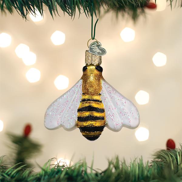 A glass bee ornament with glittery wings and gold and black stripes hangs from a Christmas tree branch, with blurred festive lights in the background.