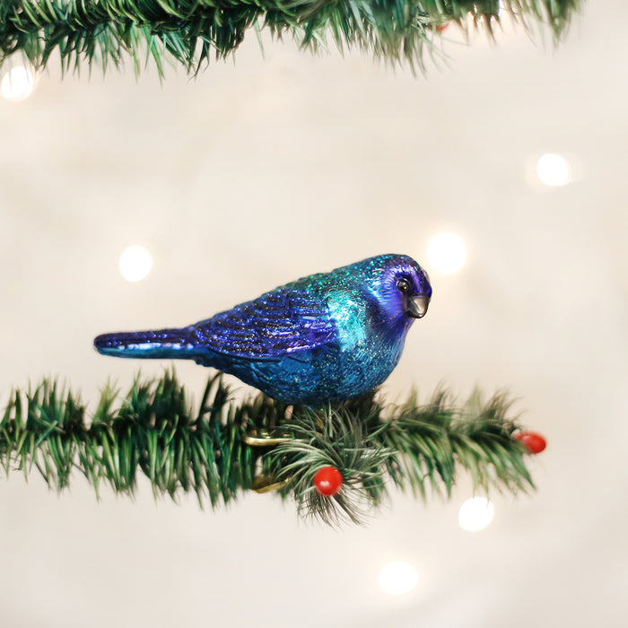 A shiny blue and purple bird ornament is perched on a green artificial pine branch with red berries, against a soft, blurred background of white lights.