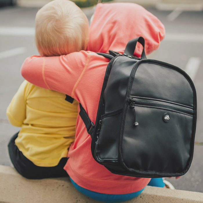 Lifestyle photo of a two little kids. Once is wearing the small black backpack while hugging the smaller little brother in the image