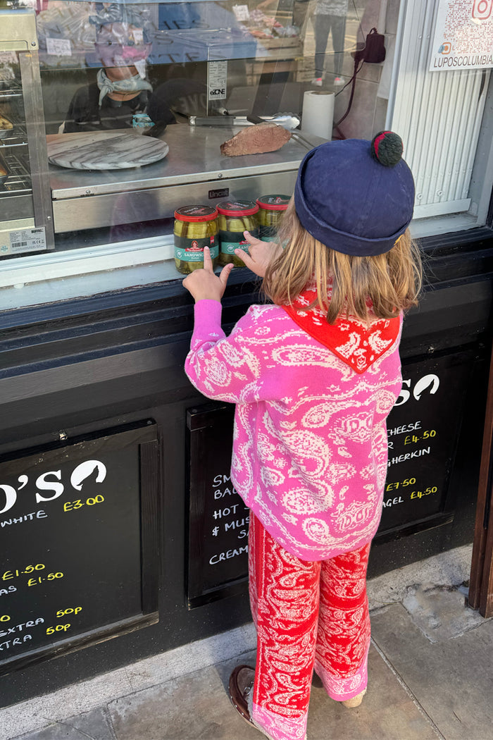 Kids Red and Pink Bandana Knitted Set featuring a paisley print jumper and trousers, worn by a child at a shop window.