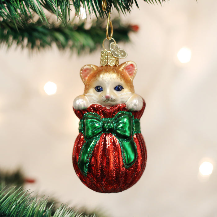 A Christmas tree ornament shaped like a kitten peeking out of a shiny red stocking with a large green bow, hanging from a branch with blurred holiday lights in the background.
