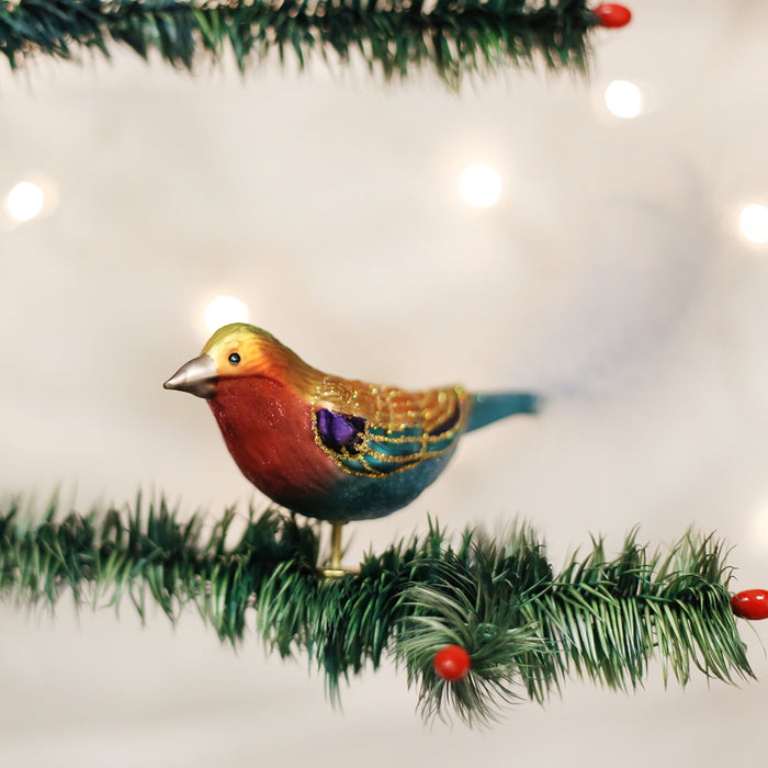 A colorful bird ornament with a red chest and gold accents perches on a green artificial pine branch, decorated with small red berries. Soft, out-of-focus lights glow in the background.
