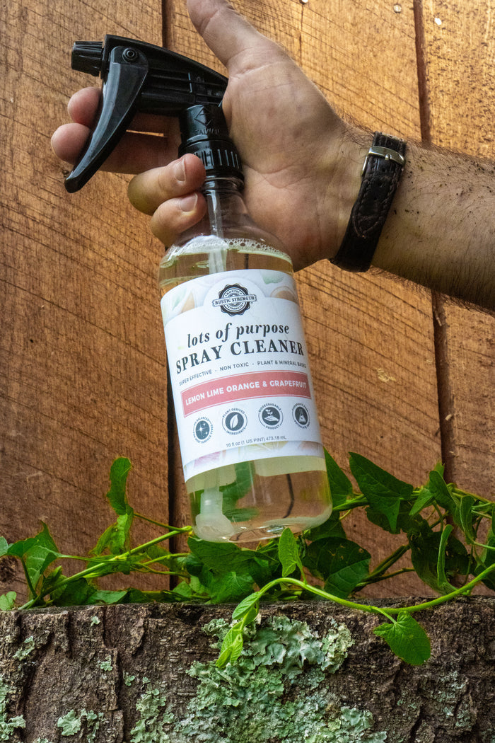 Rustic Strength, Lots of Purpose Spray Cleaner, 16oz Spray Bottle, Lifestyle image of a hand holding the bottle in a nature setting with a wooden fence background