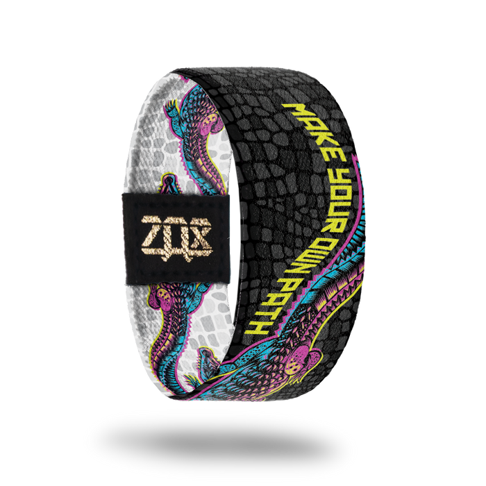 Inside product photo of Make your Own path. An alligator that is pink, yellow and blue  is shown moving its tail and the words Make Your Own Path flow with the tail. The background is black alligator scales.