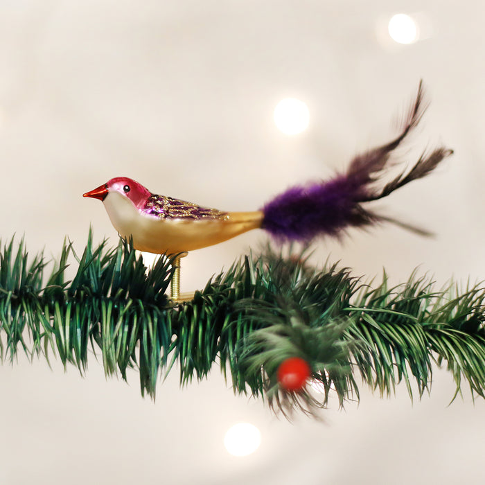 A decorative bird ornament with a purple feather tail and red beak perched on an artificial pine branch, with soft white lights blurred in the background.