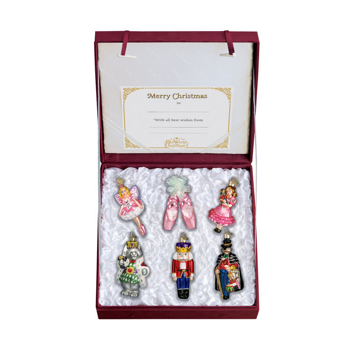 A red box with six glass Christmas ornamentsâ€”two ballerinas, a pair of pink ballet slippers, a mouse king, a nutcracker, and a soldierâ€”resting on white fabric, with a greeting card that reads Merry Christmas.