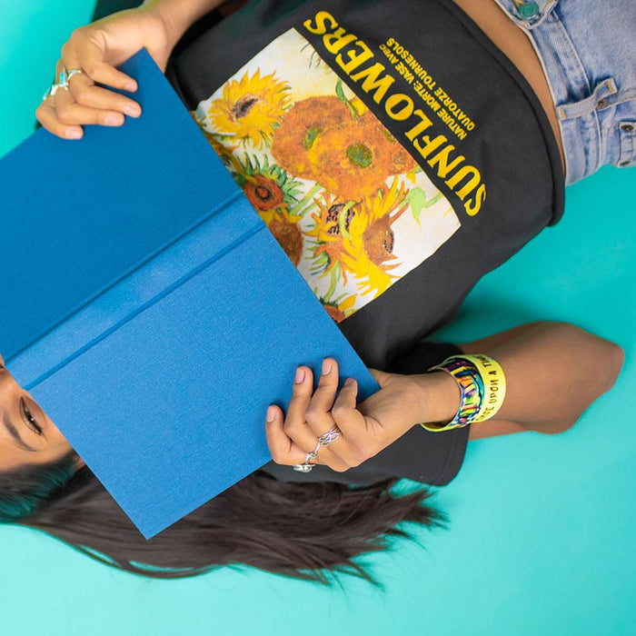 Studio image of model laying down with a blue book covering their face and 2 Once Upon A Time on their wrist