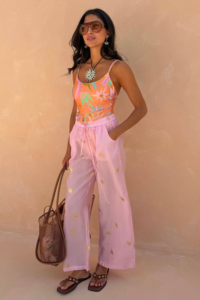 NFD Orange Dreamer Scoop Swimsuit with palm and sun prints and buckle detail, styled with pink sun-embroidered trousers.