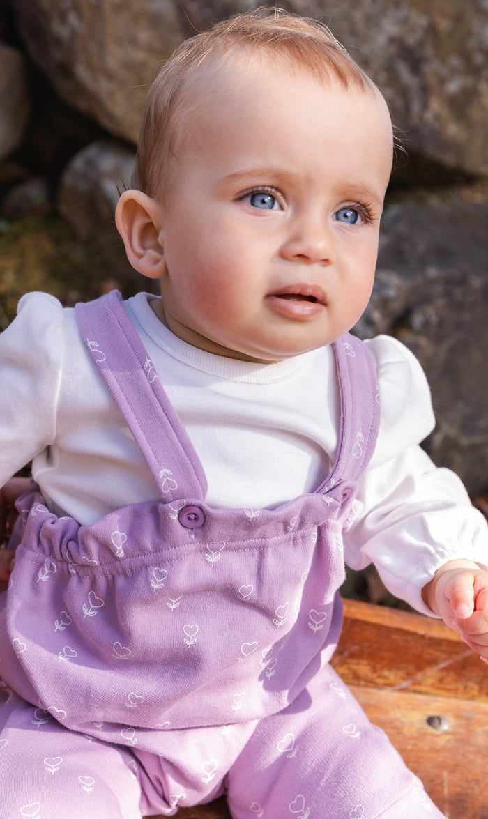 Baby wearing the Deux par Deux organic cotton onesie and purple floral overalls set, view 2