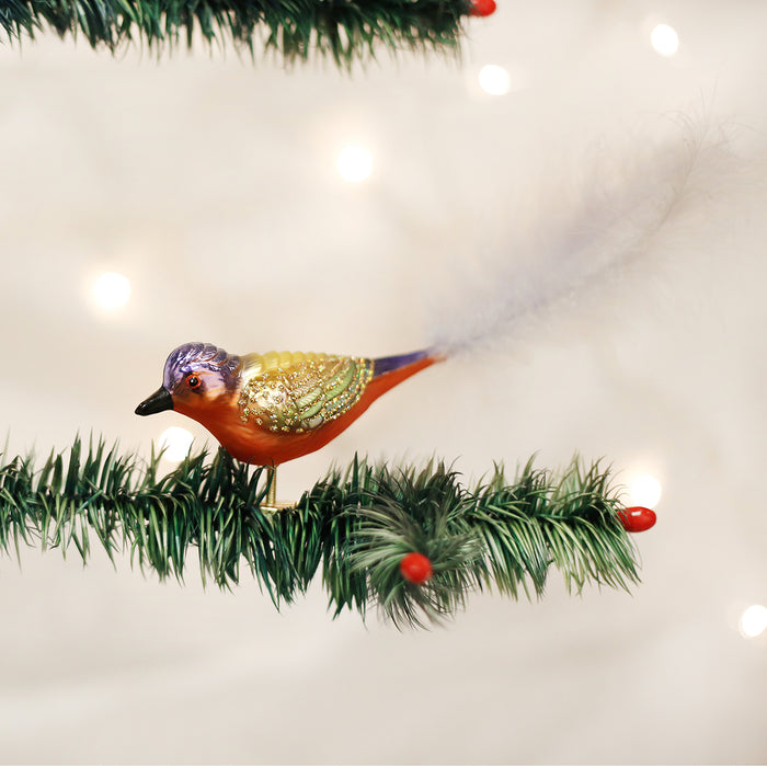 A colorful bird ornament with a long, feathery tail is clipped to a green artificial pine branch decorated with red berries, set against a background of soft, glowing holiday lights.