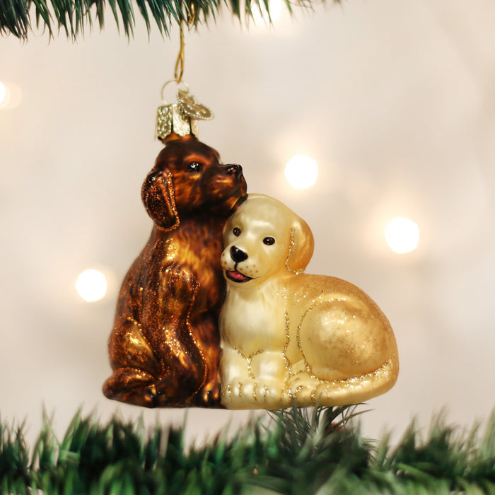 Two glass Christmas tree ornaments shaped like puppies, one brown and one light yellow, sit together on a green tree branch with warm white lights softly glowing in the background.