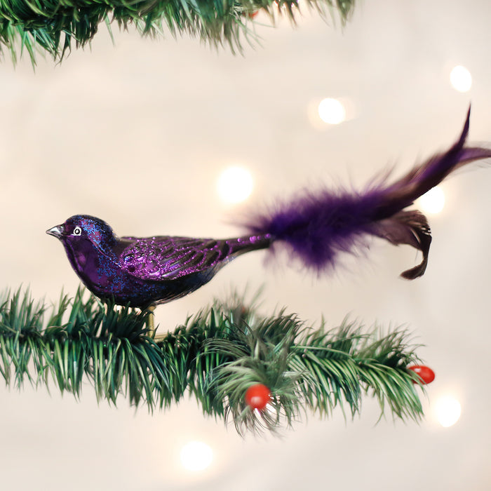 A purple bird ornament with a long feathered tail sits on a green pine branch with red berries, set against a softly blurred background with white lights.