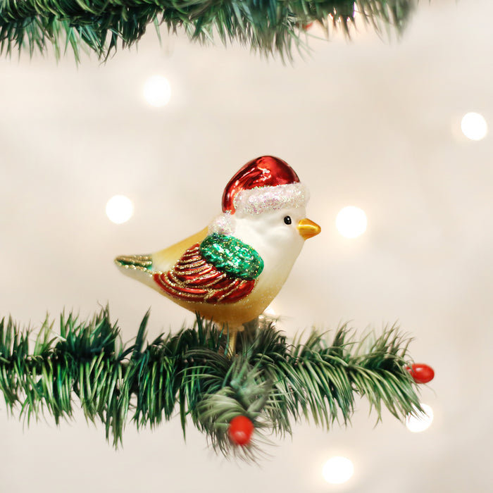 A bird-shaped Christmas ornament with a red Santa hat sits on an artificial green pine branch, decorated with red berries, against a blurred background of white holiday lights.