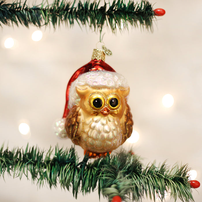 A gold and white owl ornament wearing a red Santa hat hangs from a green Christmas tree branch with blurred lights in the background.